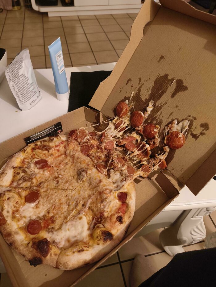 An open pizza box with half the pizza toppings, including meatballs and pepperoni, slid to one side, a food delivery fail.