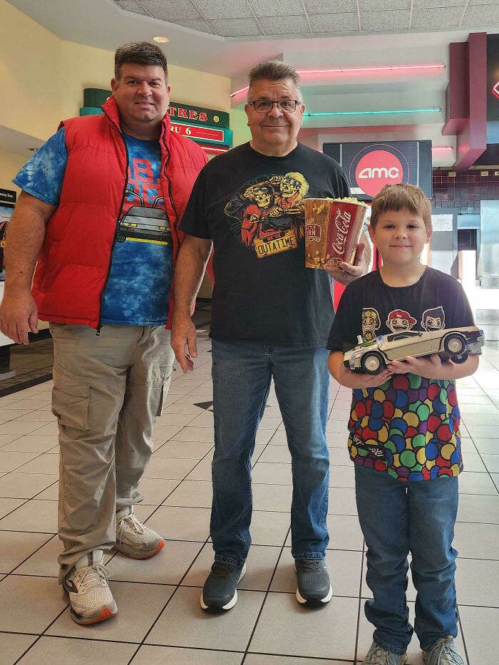 Amazing fathers: A father and two sons stand in a movie theater lobby, one holding popcorn, the other a toy car, representing amazing parents.