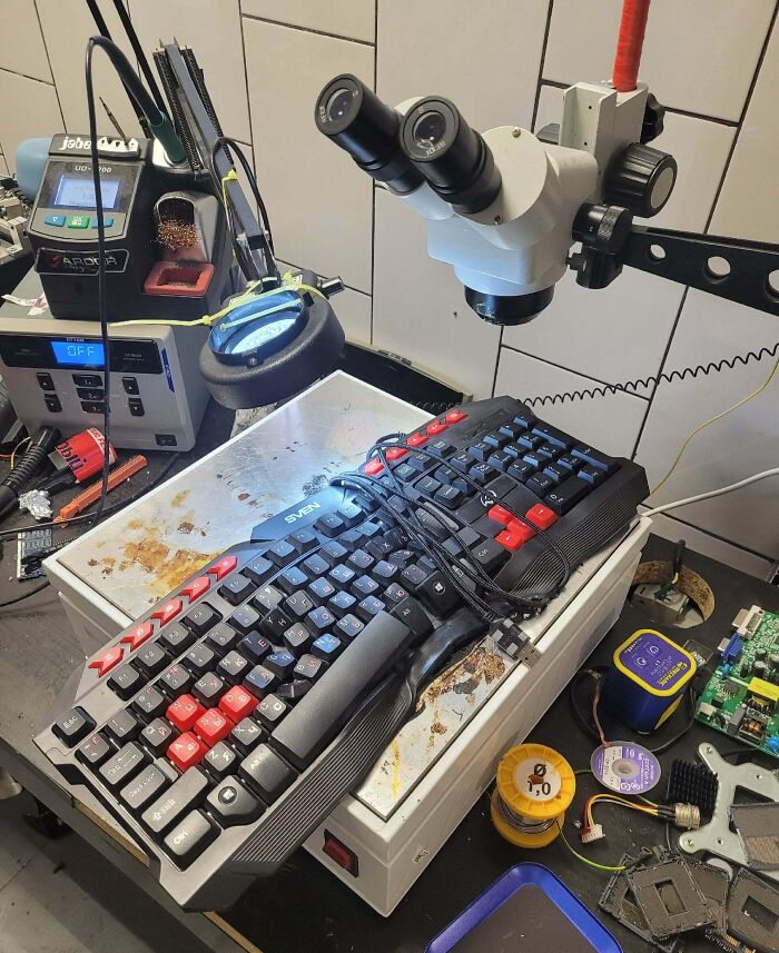 A work desk with a microscope overlooking a gaming keyboard tied with wires, showcasing Electronics Fails and the IT Guy's work.