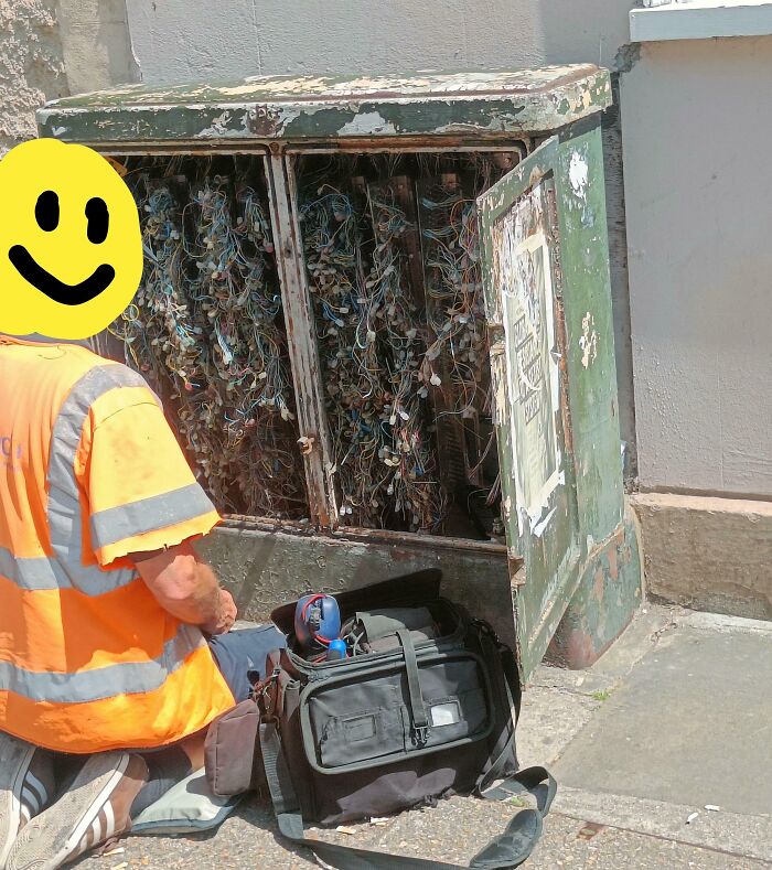A utility worker fixing a severely damaged and exposed outdoor electrical cabinet filled with tangled wires, depicting major electronics fails.