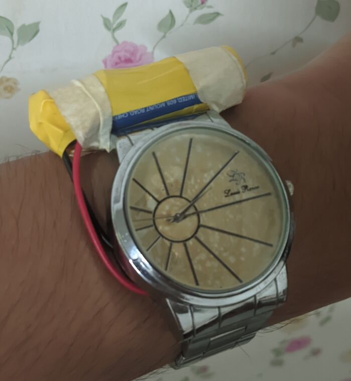 A wrist watch with a large yellow battery taped to its side, demonstrating electronics fails.