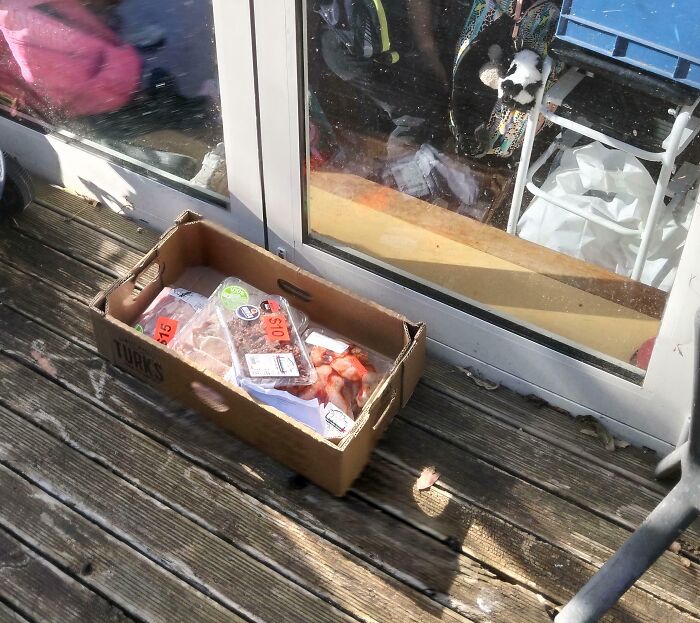 A cardboard box filled with packaged meat and strawberries sits on a wooden deck next to a glass door, a food delivery fail.