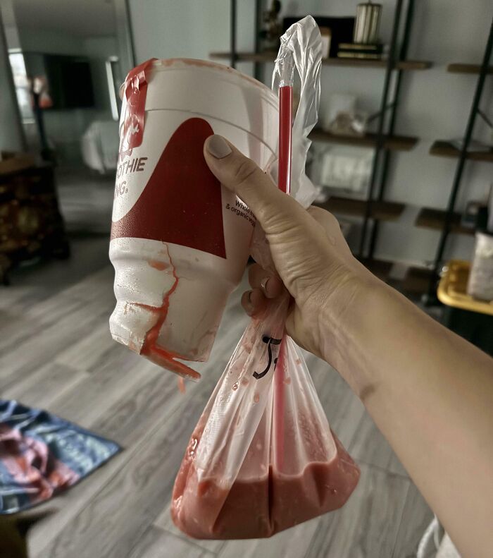 A hand holds a smoothie cup that has spilled most of its contents into a clear plastic bag, a food delivery fail.