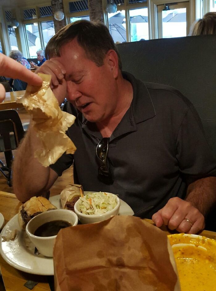 A father looking distressed and holding his head, with plates of food in front of him, at a restaurant.