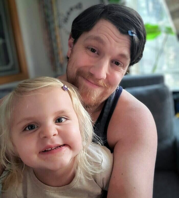 A loving father with bobby pins in his hair, hugging his smiling young daughter with blonde hair.