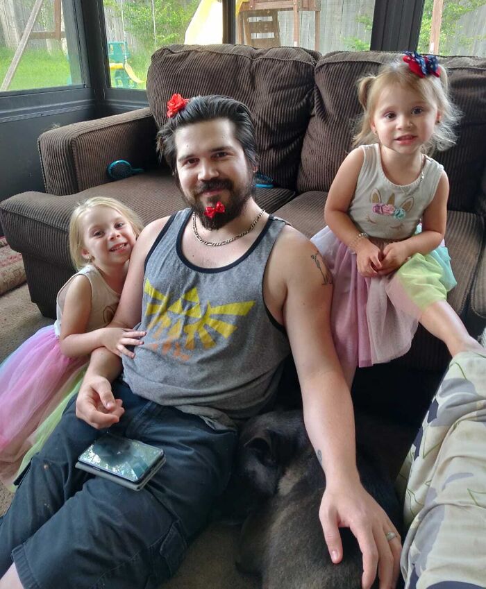 An amazing father with two young daughters, all wearing bows, sitting on a couch. The father has a small red bow in his beard.