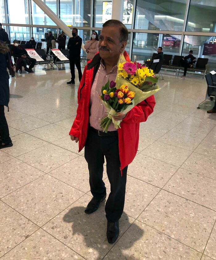 A happy father holding two bouquets of flowers, yellow and pink, in an airport terminal.