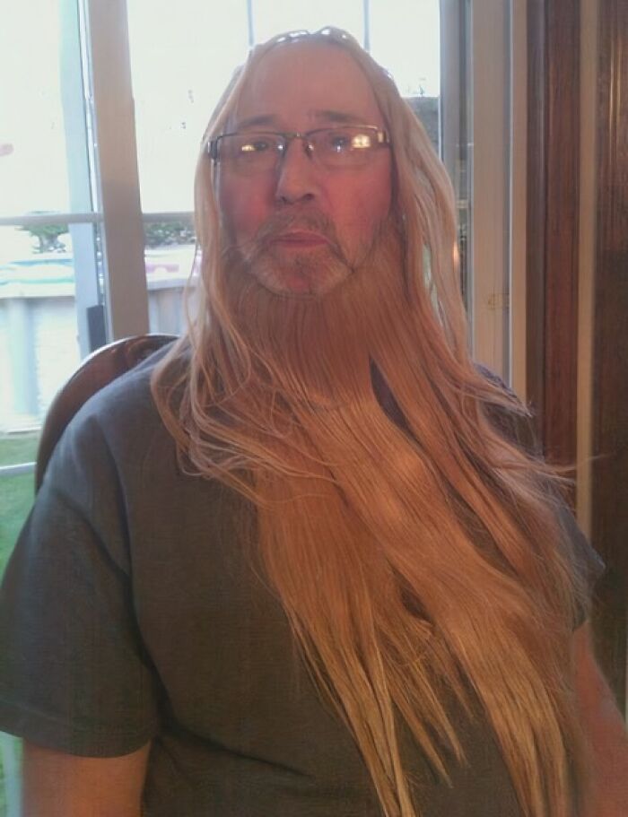 A father wearing glasses with long blonde hair draped over his head and face, creating a humorous beard.