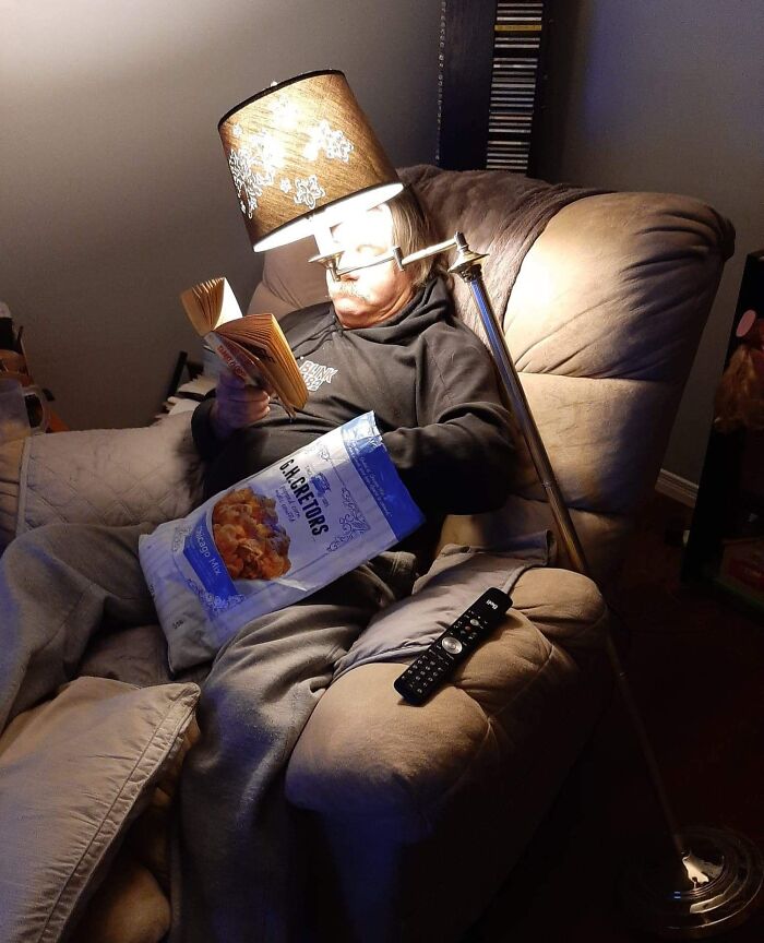 A man, an amazing father, reads a book with a lampshade on his head for light, displaying his parenting skills.