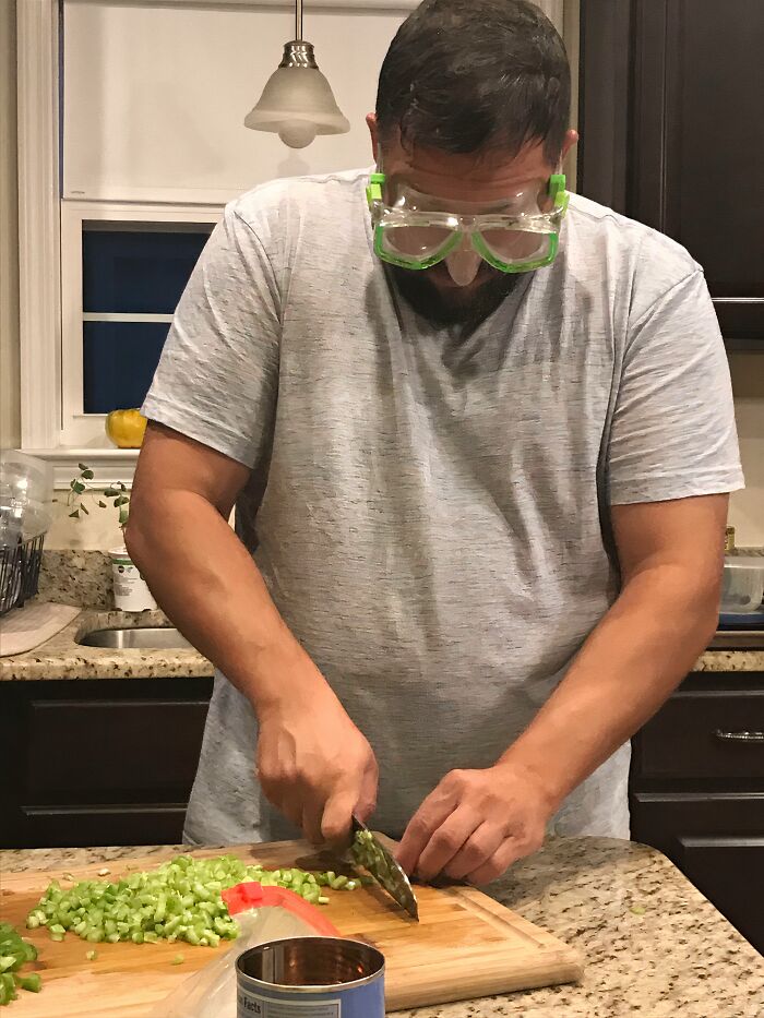 An amazing father chops vegetables in a kitchen while wearing swim goggles, demonstrating his unique parenting style.
