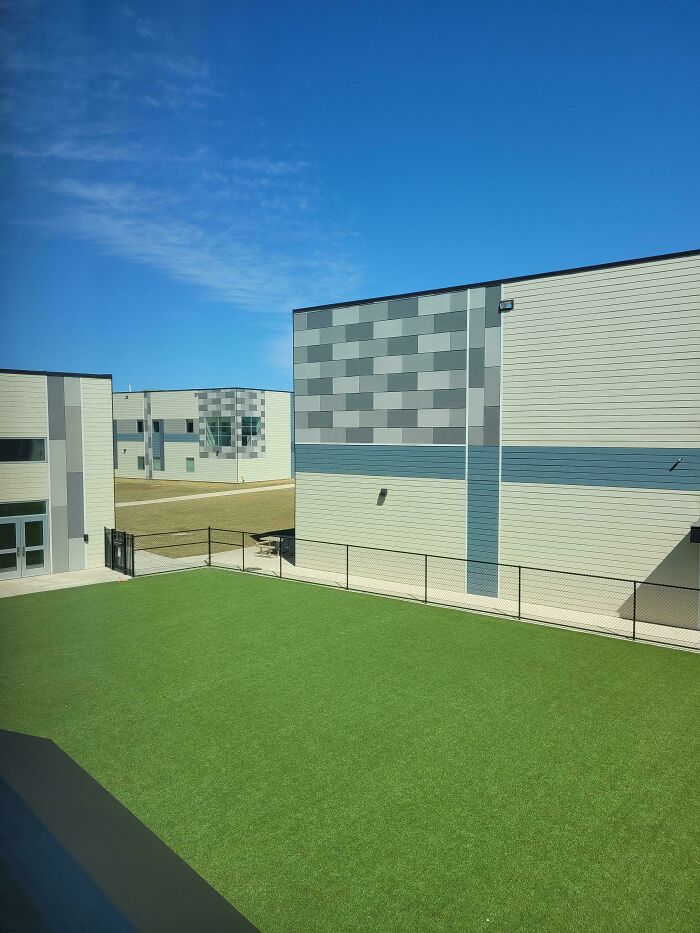 Modern buildings with a large green artificial turf area under a bright blue sky, an unsettling liminal space.