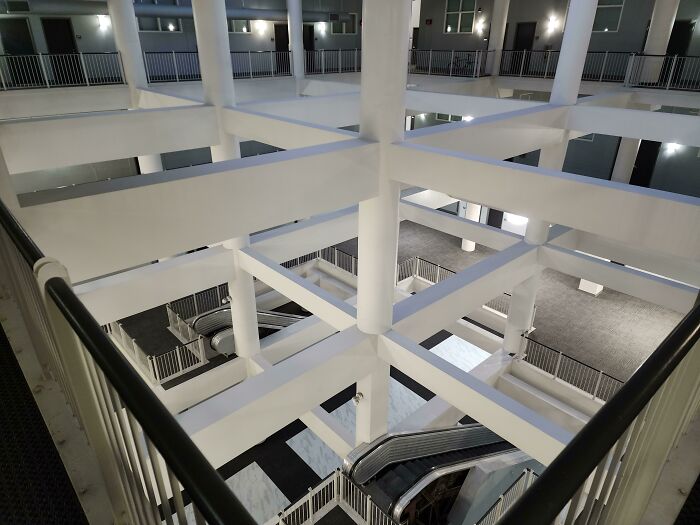 An unsettling interior view of a multi-story building with white beams and escalators, showcasing a liminal space.