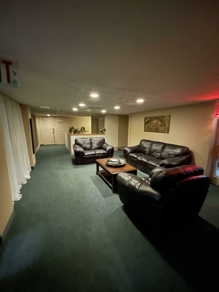 An unsettling liminal space featuring a dimly lit, empty lounge area with dark leather sofas and a green carpet, evoking an abyss.