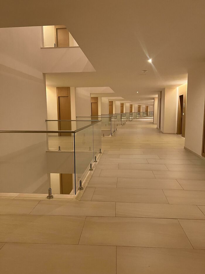 A long, deserted modern hallway with glass railings and many doors, an unsettling liminal space.
