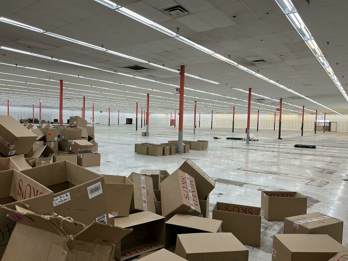 An empty, vast warehouse floor scattered with cardboard boxes, presenting a desolate and unsettling liminal space.