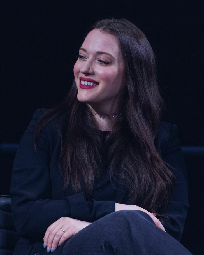 Kat Dennings smiling during an event in a black outfit with long dark hair