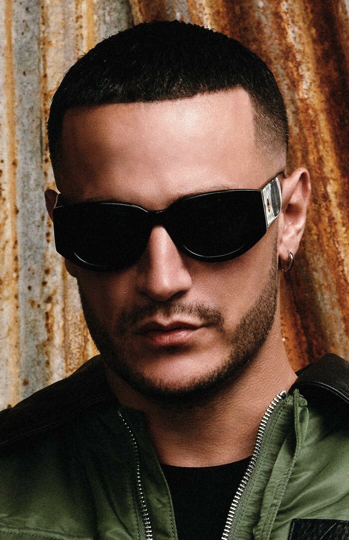 Close-up portrait of DJ Snake wearing dark sunglasses and green jacket with a textured background