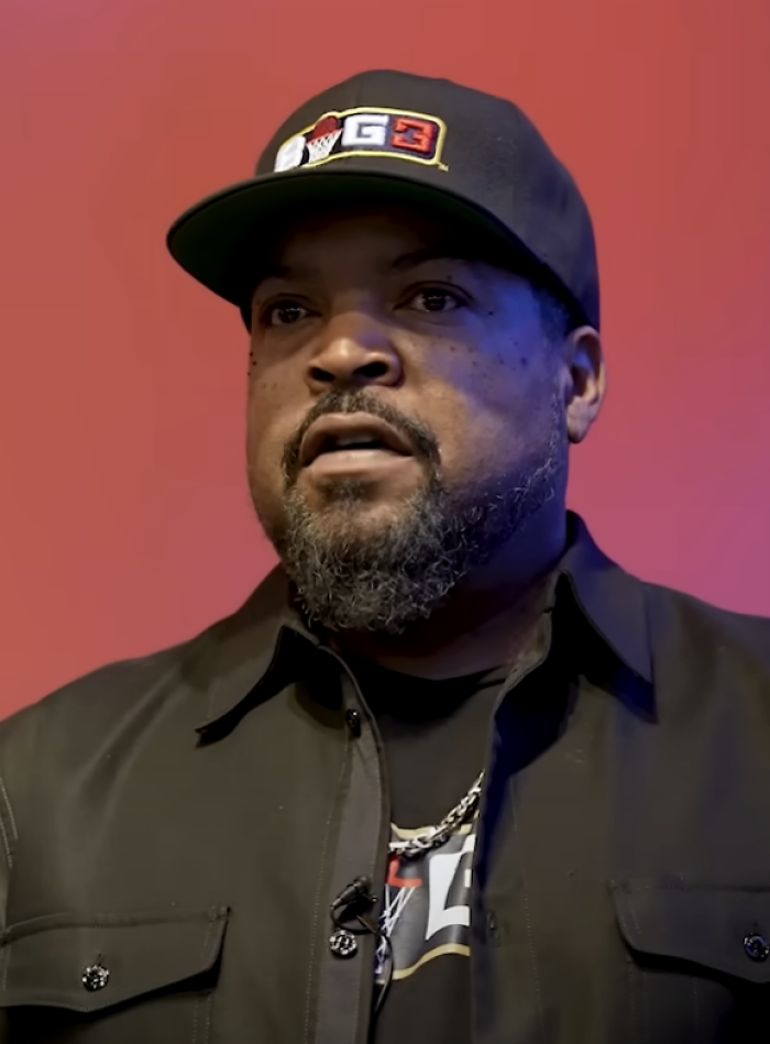 Portrait of rapper and actor Ice Cube wearing a G3 baseball cap and black shirt against a red background