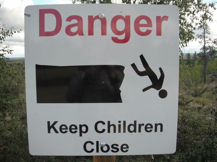 A white danger sign with red text and a black pictogram showing a person falling off a cliff, warning to keep children close.