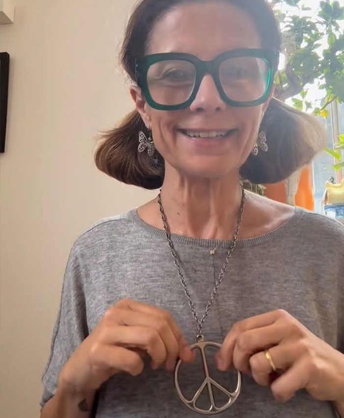 Smiling woman with large glasses holding peace sign necklace indoors Smiling woman with large glasses holding peace sign necklace indoors