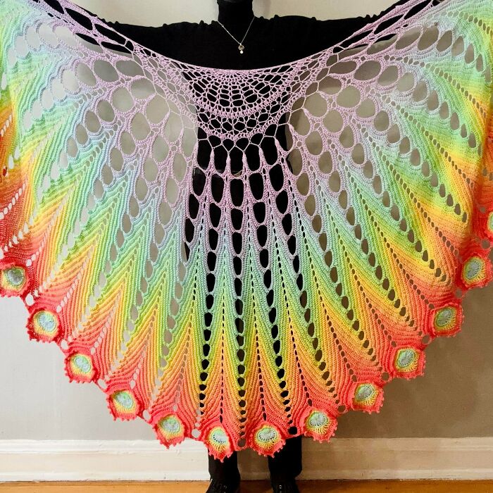 A person holding up a large handmade crochet shawl in rainbow colors, showcasing incredible talent and patience.