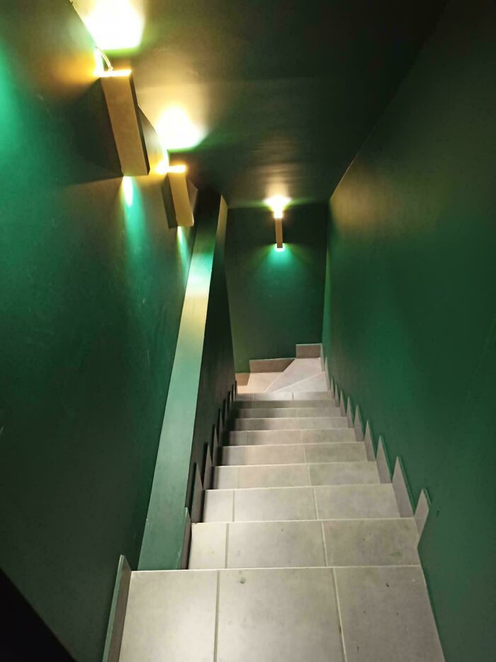A narrow stairway with green walls and bright wall sconces, descending into an unsettling liminal space.