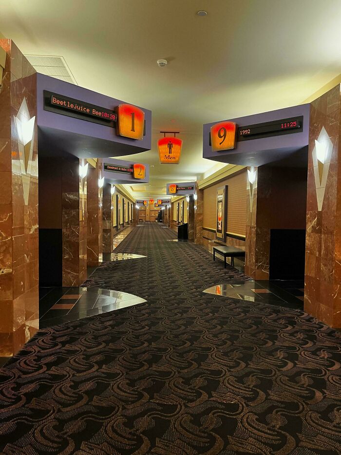 A long, empty movie theater hallway with patterned carpet and numbered signs for screens, an example of a liminal space.