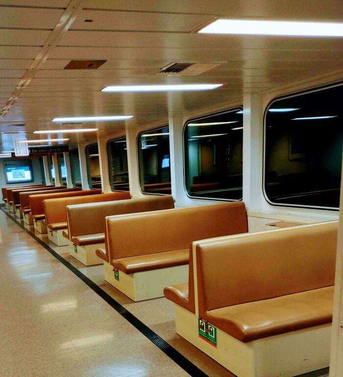 An empty ferry cabin with rows of brown benches and large windows, illustrating an unsettling liminal space.