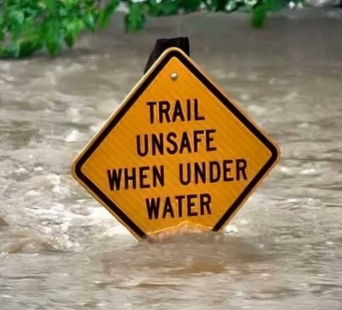 A funny sign in the wild that says Trail Unsafe When Under Water, partially submerged in floodwaters.