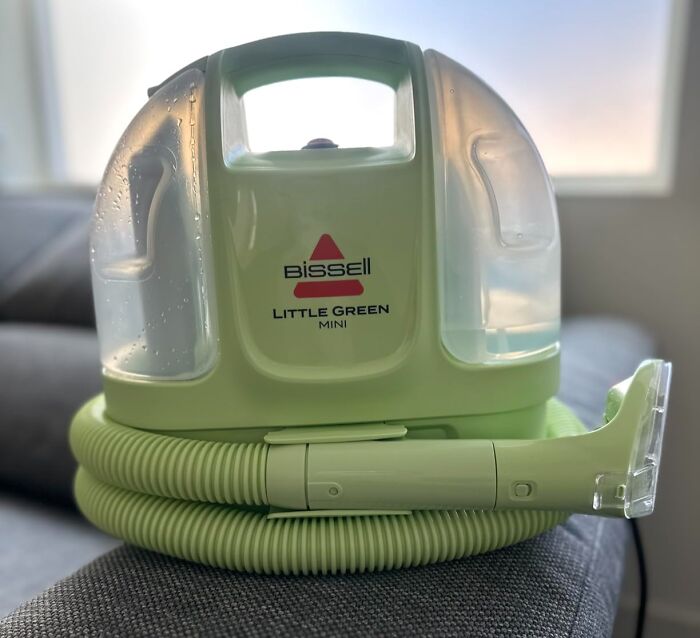A green Bissell Little Green Mini portable carpet cleaner on a grey sofa. Don't miss these Prime Day Deals on home cleaning.