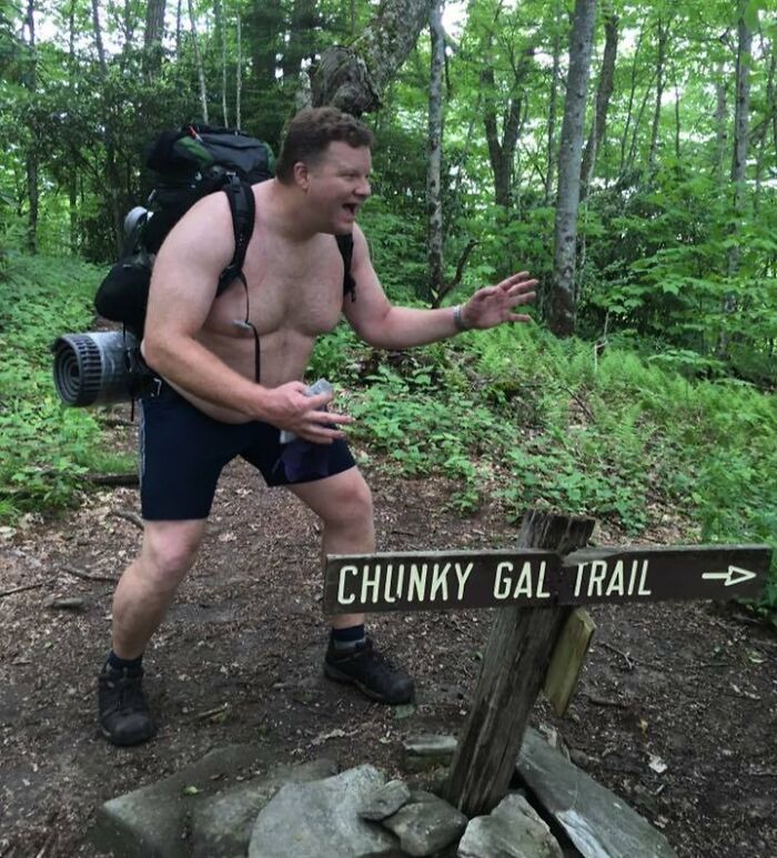A man with a backpack posing next to a funny sign pointing to Chunky Gal Trail in a forest.