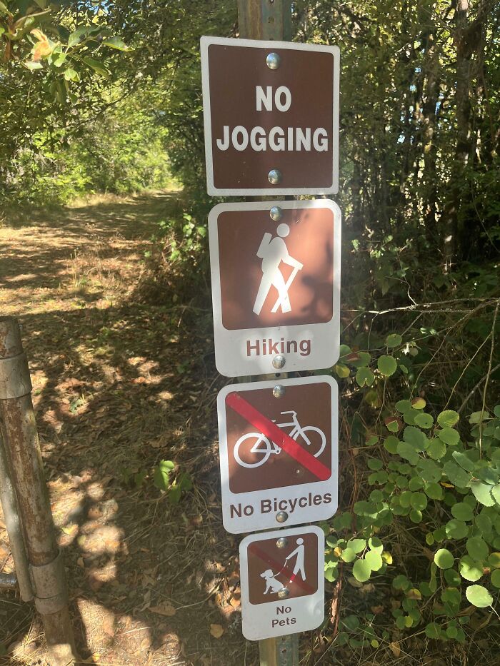 A brown and white signpost with multiple funny signs: No Jogging, Hiking, No Bicycles, and No Pets allowed on the trail.