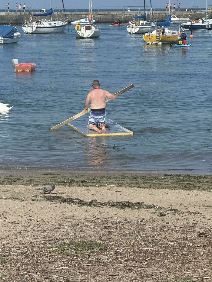 A mad lad on a homemade raft paddling with a wooden plank in a harbor full of boats, displaying a personal challenge.