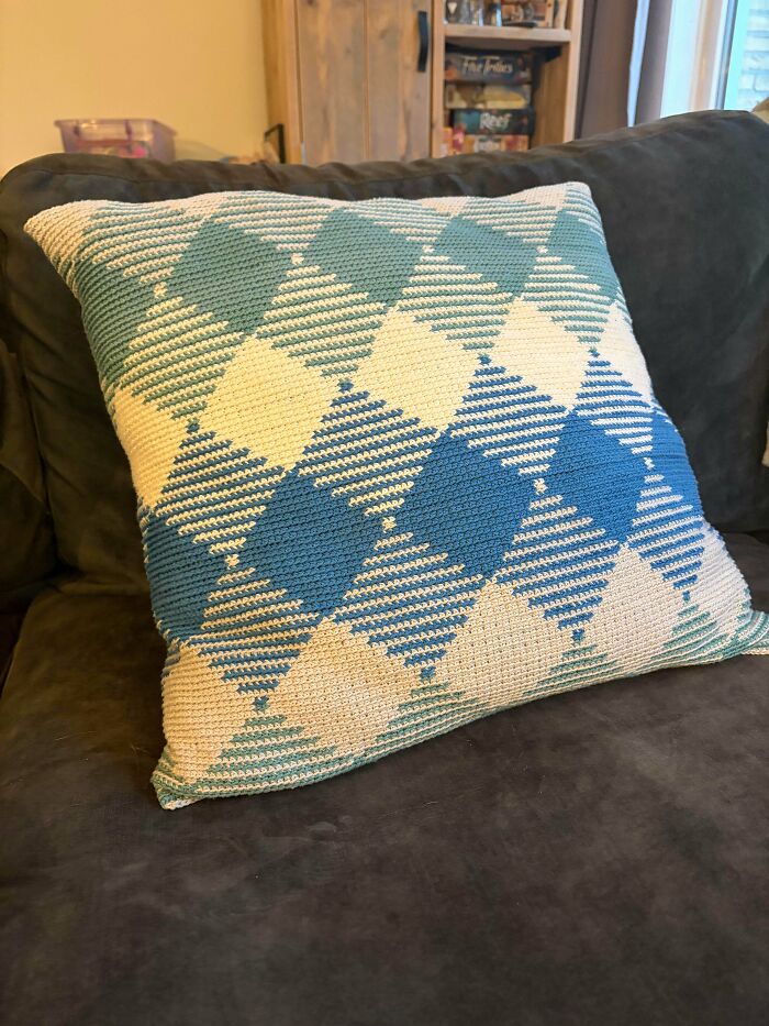 A crocheted throw pillow with a geometric blue and white diamond pattern rests on a dark gray couch, a stunning piece by crochet enthusiasts.