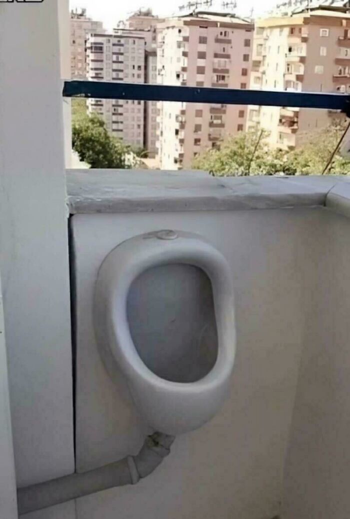 A urinal installed on an outdoor balcony of an unhinged real estate listing, overlooking city buildings.