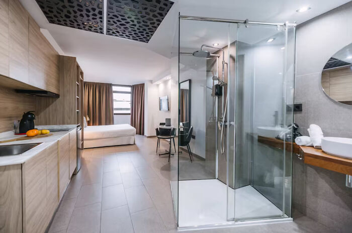 An unhinged real estate listing featuring an open-concept studio apartment with a bed, kitchen, and glass shower.