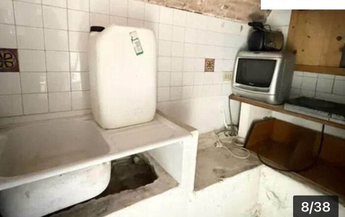 A small, grimy utility room with an old sink, a large white plastic container, and an outdated TV, part of unhinged real estate listings.
