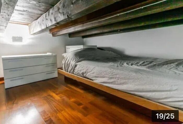 Unhinged real estate listings showcasing a bed crammed under low wooden beams, creating an uncomfortable and claustrophobic sleeping space.