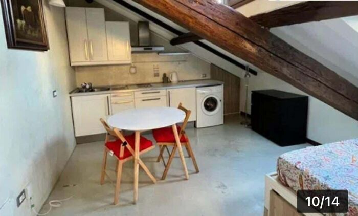 An unhinged real estate listing featuring a small kitchen and dining area under sloped ceiling beams, with a bed just out of frame.