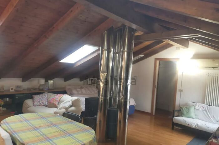 Unhinged real estate listing: an attic living space with exposed wooden beams and a large, shiny metal chimney pipe dominating the center.