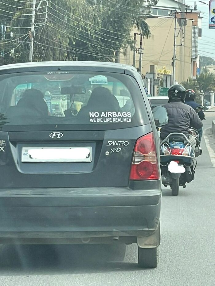 A car with a sticker on the back window saying NO AIRBAGS WE D*E LIKE REAL MEN, demonstrating toxic masculinity.