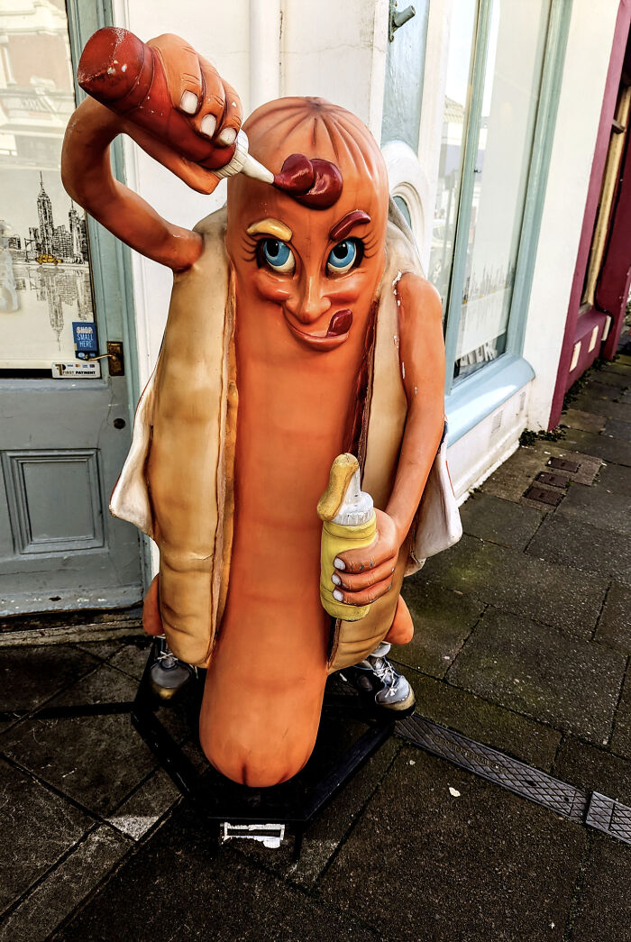 A creepy hot dog design fail statue outside, squeezing ketchup and mustard onto itself with a strange expression.