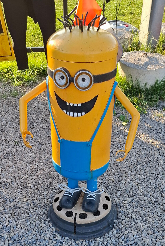 A Minion made from yellow metal barrels with large goggles, a creepy design fail in a yard.