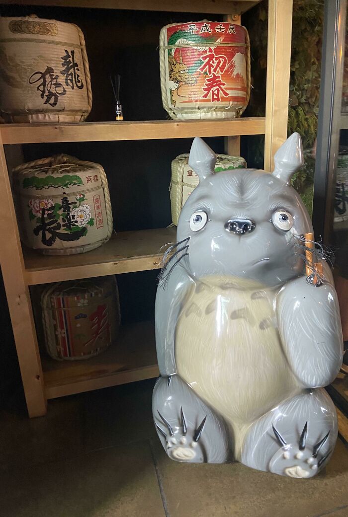 A gray Totoro statue holding an item, a creepy design fail displayed on a shelf with sake barrels.