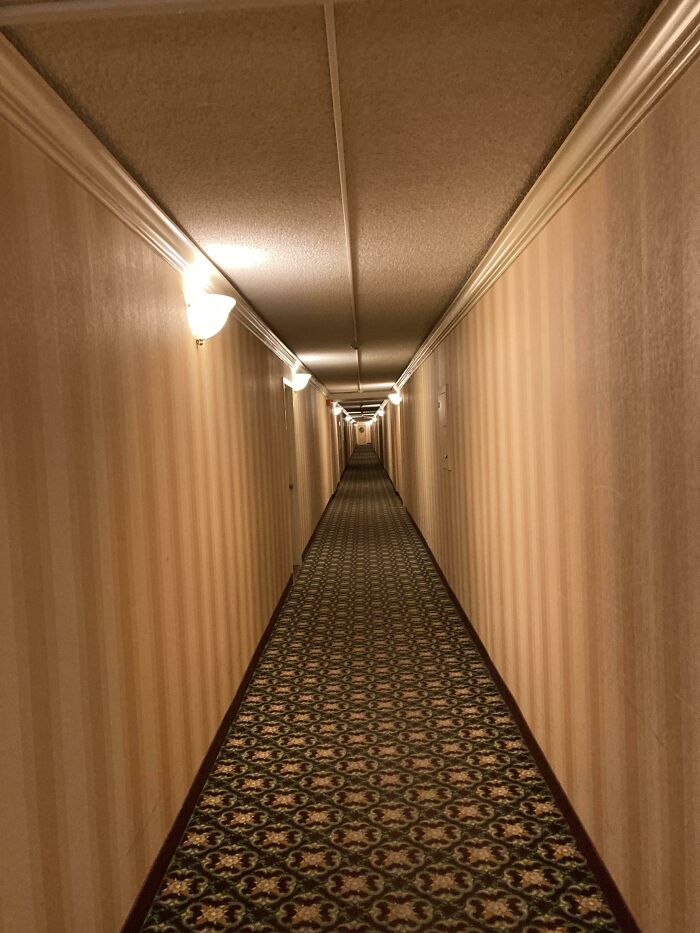 A creepy, long, dimly lit hotel hallway with patterned carpet and striped wallpaper, a design fail.