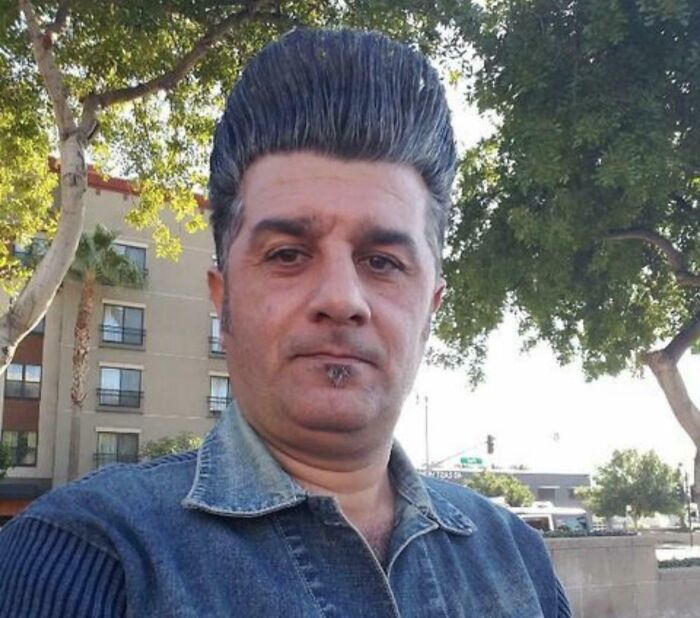 Man with a very tall, unfortunate haircut, wearing a denim shirt outdoors.