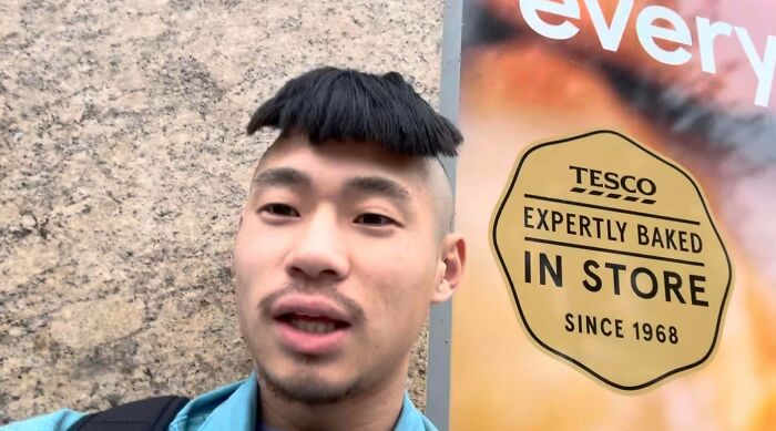 A man with an unusual and unfortunate haircut, featuring a short, blunt fringe over his forehead, standing near a Tesco sign.