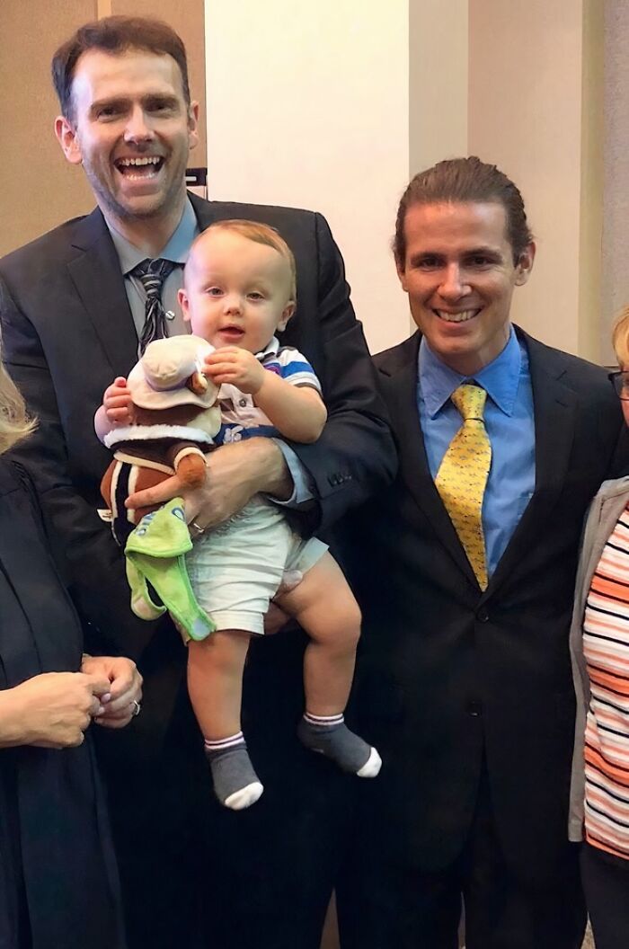 A happy family celebrating their adoption story with a baby held by a smiling man in a suit.
