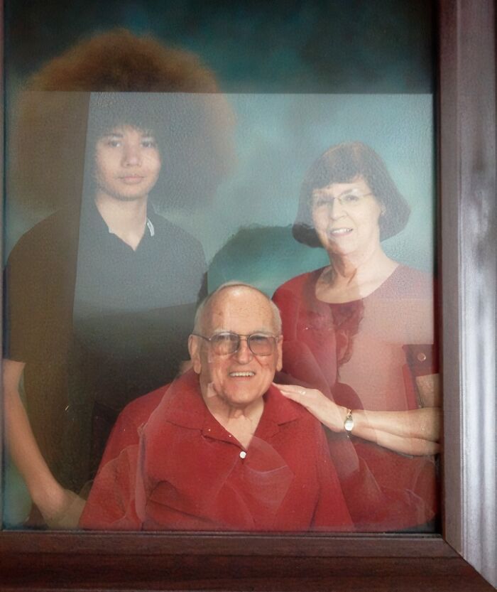 A framed photograph of a family of three: an older man sitting, an older woman standing with her hand on his shoulder, and a young man standing behind, illustrating adoption stories.
