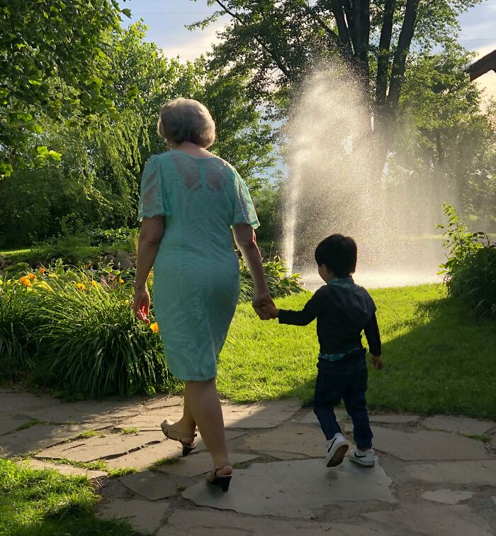 An older woman in a blue dress walks hand-in-hand with a young boy through a garden, showing adoption stories.